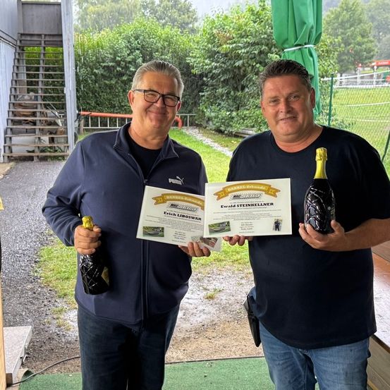 Zwei Männer stehen im Freien, lächeln und halten eine Flasche Champagner und ein Zertifikat. Sie tragen Freizeitkleidung, wobei einer der Männer ein Zertifikat mit dem Namen Erich Libowar hält und der andere ein Zertifikat mit dem Namen Ewald Steinkellner. Hinter ihnen befindet sich ein Rasenbereich mit einem Zaun.