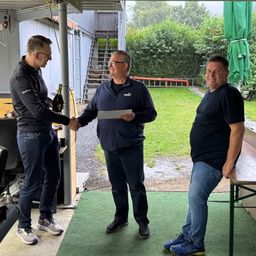 Drei Männer stehen in einem Schuppen; einer reicht einem anderen ein Papier und hält eine Flasche. Hinter ihnen ist ein grüner Schirm, eine Treppe und ein Rasen.