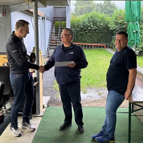 Drei Männer stehen in einem Schuppen; einer reicht einem anderen ein Papier und hält eine Flasche. Hinter ihnen ist ein grüner Schirm, eine Treppe und ein Rasen.