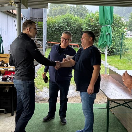 Drei Männer schütteln sich in einem überdachten Außenbereich die Hände. Ein Mann hält Stift und Papier. Ein anderer Mann hält eine Flasche. Eine Bank und ein grüner Regenschirm sind im Hintergrund.