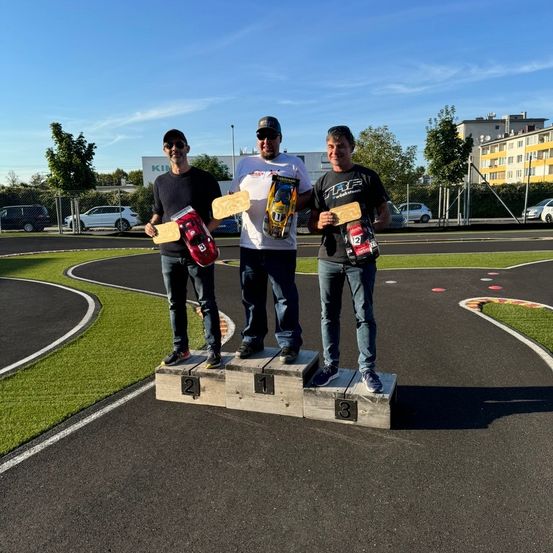 Drei Männer stehen auf einem Podest und halten Trophäen und Modellautos. Sie sind auf einer Rennstrecke mit einem Rasenbereich und einem Zaun. Dahinter sind geparkte Autos und ein Gebäude.