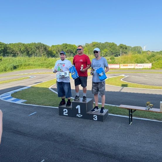 Drei Männer stehen auf einem Podium auf einer Rennstrecke. Sie halten Pokale und ein kleines Automodell. Das Podium hat die Zahlen 1, 2 und 3. Dahinter befindet sich ein Rasenbereich mit Bäumen.
