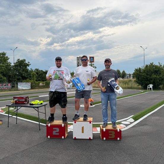 Drei Männer stehen auf Podestplätzen einer Rennstrecke, halten Trophäen und Spielzeugautos. Die Strecke hat Straßenlaternen und Banner. Bäume und ein bewölkter Himmel im Hintergrund.