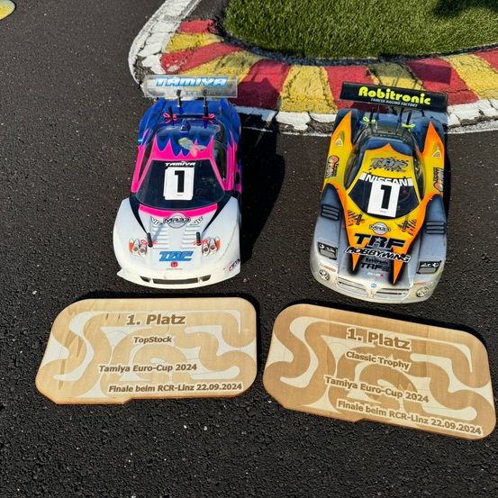 Zwei Tamiya-Rennwagen, einer weiß mit rosa Design und der andere gelb, stehen auf der Rennstrecke. Beide haben die Nummer 1 auf der Vorderseite. Unter jedem Wagen befindet sich eine Holzplakette mit '1. Platz' und einer Erwähnung des Tamiya Euro-Cup 2024.