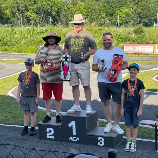 Drei Männer und zwei Jungen stehen auf einem Podest. Die Männer tragen Hüte und halten Trophäen. Die Jungen tragen Medaillen. Sie sind auf einer Rennstrecke mit einem Rasenbereich und einem Zaun. Bäume und ein Gebäude sind im Hintergrund.