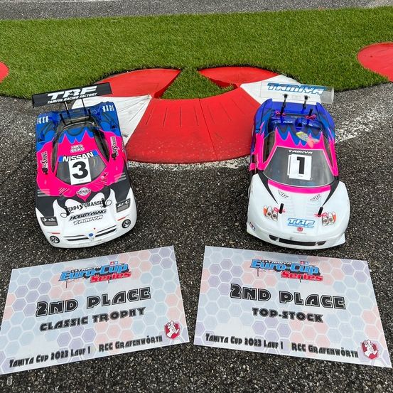 Zwei ferngesteuerte Rennwagen, einer pink und schwarz mit der Nummer 3 und der andere pink und weiß mit der Nummer 1, sind auf einer Rennstrecke ausgestellt. Sie stehen neben zwei Trophäen, eine mit der Aufschrift 2. Platz Classic Trophy und die andere 2. Platz Top-Stock.