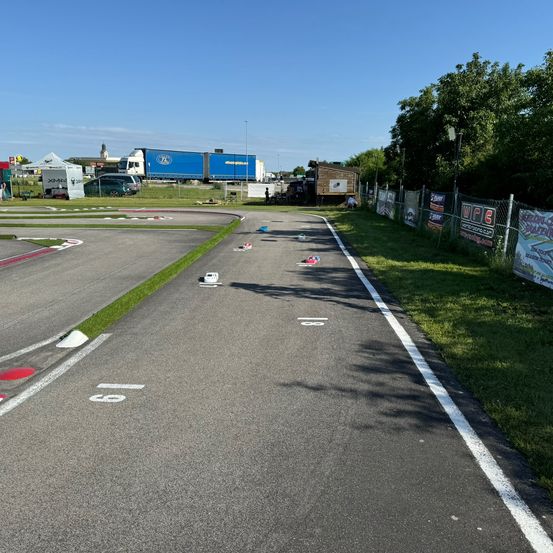 Eine leere Rennstrecke mit einer weißen Spurlinie und kleinen Autos in der Ferne. Es gibt Zelte und geparkte Autos an der Seite.