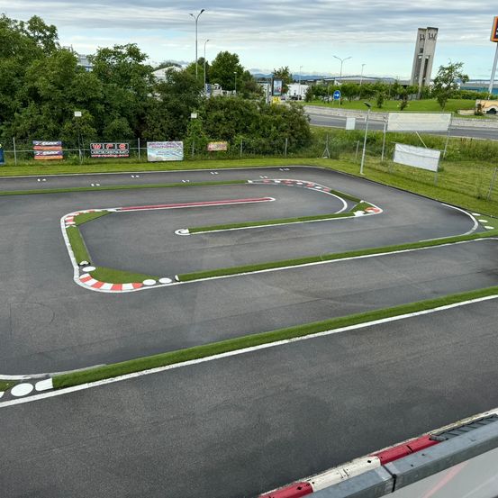 Eine Rennstrecke mit einer kurvigen Anordnung und grasbewachsenen Bereichen an den Seiten. Es gibt weiße und rote Markierungen auf der Strecke und Banner entlang des Zauns.