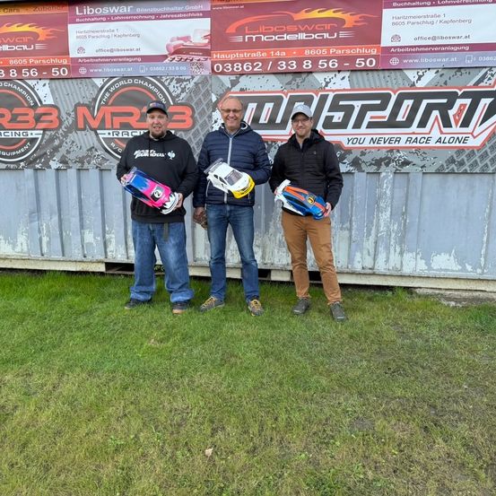 Drei Männer stehen auf einem Rasenfeld, halten Trophäen und lächeln. Hinter ihnen ist eine Wand mit Bannern, die 'Liboswar Modellbau' und 'MR3 Racing' bewerben.