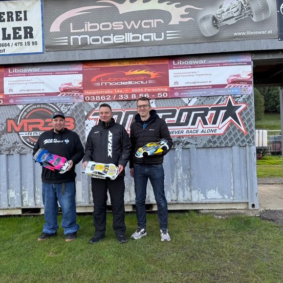 Drei Männer stehen vor einem Gebäude mit Schildern für Liboswar Modellbau und CORTX. Sie halten Rennwagen und scheinen ein Event zu feiern.