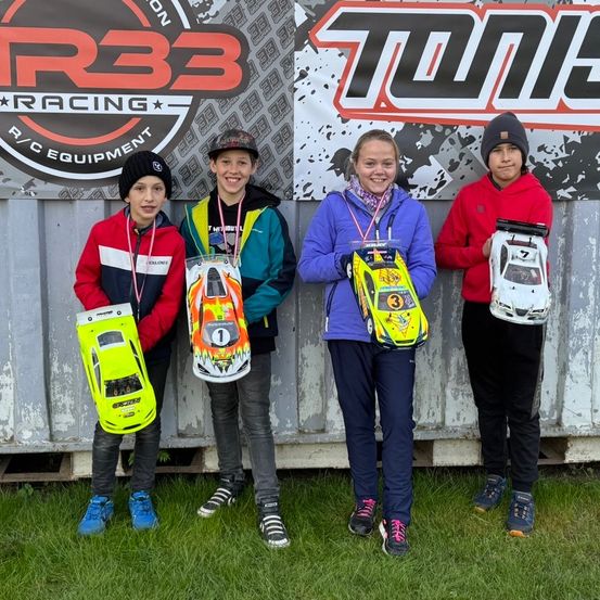 Vier Kinder in Winterkleidung posieren mit ferngesteuerten Autos in den Haenden vor einer grauen Wand. Dahinter befindet sich ein Banner mit dem Text 'TONG RACING'.