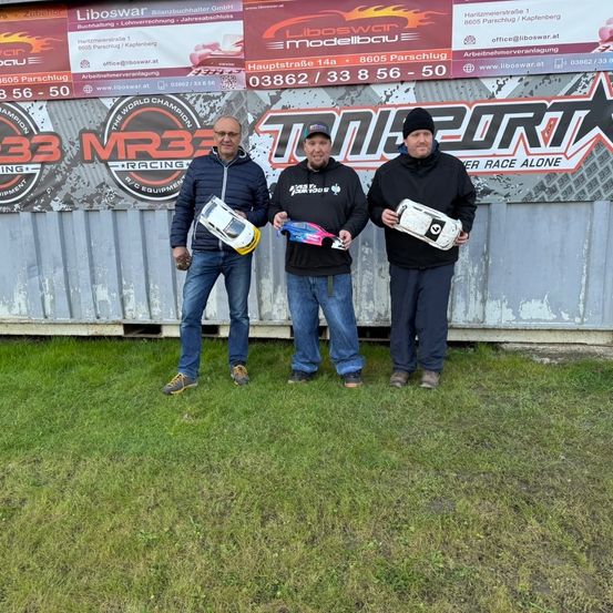 Drei Männer stehen vor einem Gebäude mit einem Banner und halten ferngesteuerte Autos in den Händen. Sie lächeln und scheinen für ein Foto zu posieren.