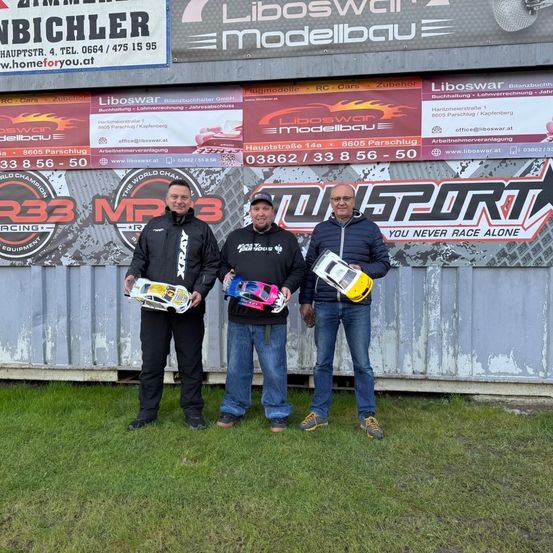 Drei Männer posieren mit ferngesteuerten Autos für ein Foto vor einer Wand mit verschiedenen Werbetafeln, darunter eine für Liboswar Modelbau.