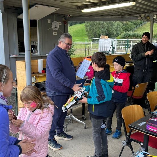 Ein erwachsener Mann gibt einem Kind in einer Gruppe ein Spielzeug. Das Kind hat rosa Farbe im Gesicht. In der Nähe sind andere Kinder und Erwachsene. Dahinter sind eine Uhr und ein Regal zu sehen.