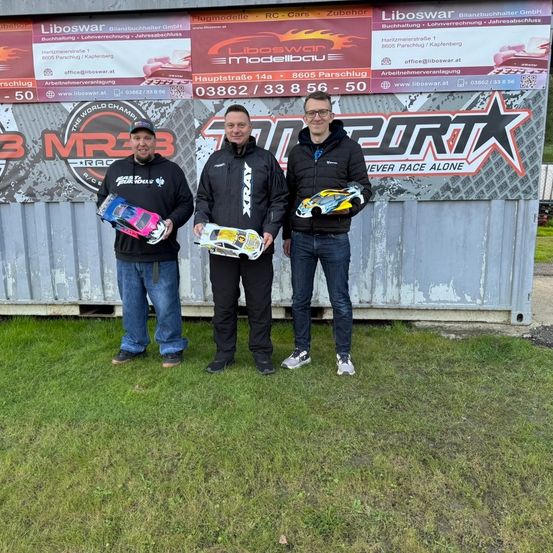 Drei Männer in Winterkleidung stehen vor einer Metallwand mit Werbung. Sie halten Miniaturrennwagen. Das Banner hinter ihnen zeigt Logos, Text und Bilder.