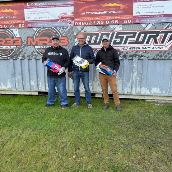 Drei Männer stehen vor einem Schild mit Logos und halten Modellautos in der Hand. Sie tragen Jacken und Jeans. Hinter ihnen befindet sich eine Metallwand mit Bannern.