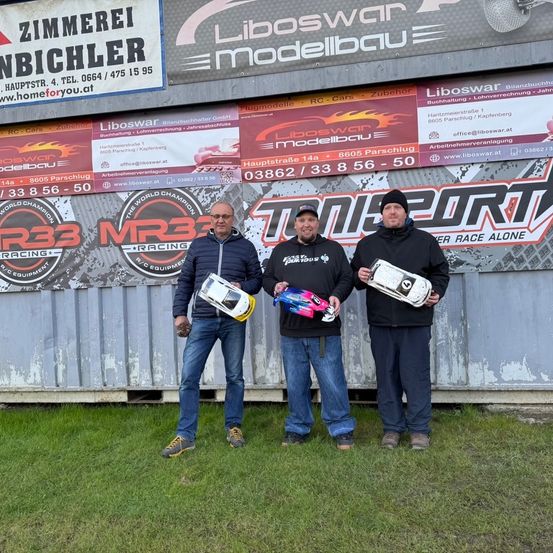 Drei Männer stehen auf Gras und halten Trophäen. Sie tragen Jacken und Jeans. Hinter ihnen zeigt ein Gebäude Schilder für Zimmerei Bichler und Liboswar Modellbau.