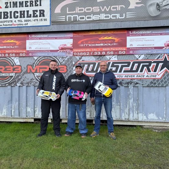 Drei Männer stehen vor einem Gebäude mit einem Banner, auf dem 'Liboswar Modellbau' steht. Sie halten ferngesteuerte Autos. Ein Mann trägt eine Brille, und ein anderer hat eine Kappe. Das Gebäude hat eine Metallwand und einen Rasenbereich.