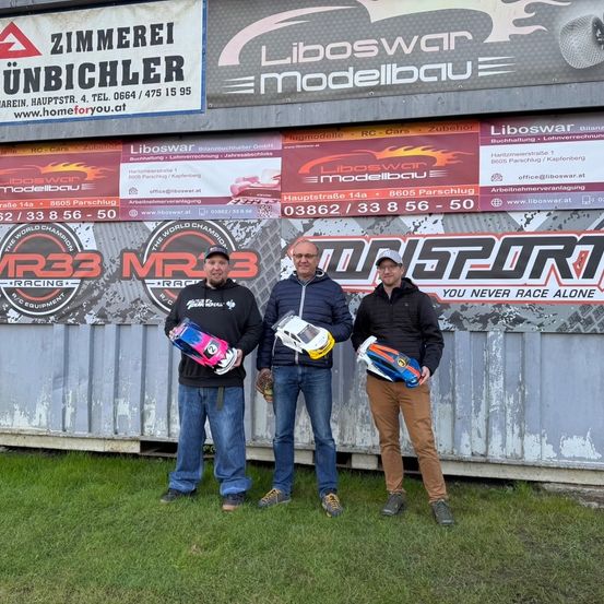 Drei Männer stehen vor einem Banner mit 'Liboswar Modellbau'. Sie halten Spielzeugautos in der Hand und posieren für ein Foto.