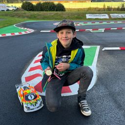 Ein junger Junge kniet auf einer Rennstrecke, lächelt und hält eine Medaille und ein Spielzeugauto. Die Strecke hat farbige Markierungen, und der Junge trägt eine Kappe und Turnschuhe.