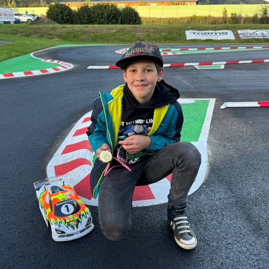 Ein junger Junge kniet auf einer Rennstrecke, lächelt und hält eine Medaille und ein Spielzeugauto. Die Strecke hat farbige Markierungen, und der Junge trägt eine Kappe und Turnschuhe.