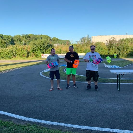 Drei Männer stehen auf einer Rennstrecke und halten ferngesteuerte Autos in den Händen. Sie tragen Freizeitkleidung und Sneakers. Hinter ihnen steht ein Tisch und ein Schild. Im Hintergrund befinden sich Bäume und ein Gebäude.