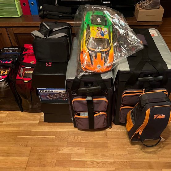 Ein Tisch mit mehreren Taschen und Kisten, auf dem ein Tamiya-Spielzeugauto steht, und verschiedene Gegenstände wie Ordner und ein Philips-Fernseher im Hintergrund.