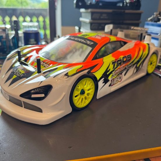 Ein weißer, orangefarbener und gelber Rennwagenmodell mit dem Wort 'TAOS' auf der Seite, auf einem Tisch stehend. Im Hintergrund befinden sich Lagerboxen und ein Glasfenster.