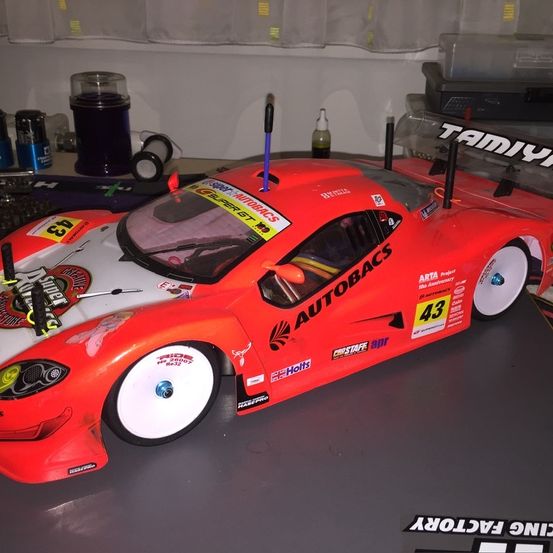 Ein rot-weißes Modellrennauto mit der Nummer 43 steht auf einer grauen Oberfläche. Es hat Antennen und mehrere Aufkleber. Dahinter befindet sich eine weiße Wand mit Behältern.