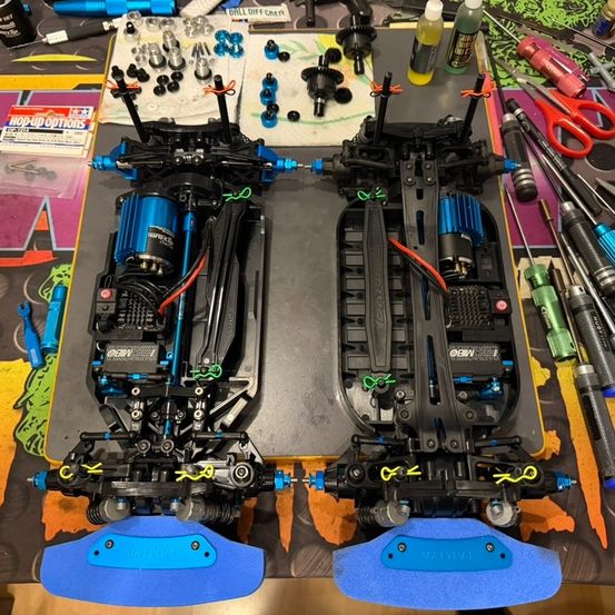 Zwei zerlegte Fahrgestelle von ferngesteuerten Autos liegen auf einem Tisch mit verschiedenen Werkzeugen und Ersatzteilen. Jedes Fahrgestell hat einen blauen Motor und sichtbare interne Mechaniken.