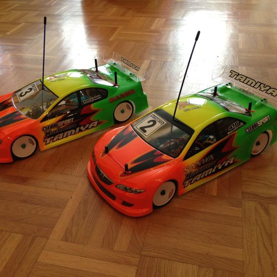 Zwei ferngesteuerte Rennwagen, einer grün und orange mit der Nummer 3 und der andere orange und grün mit der Nummer 2, stehen auf einem Holzboden.