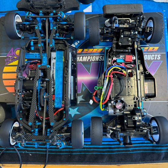 Zwei RC-Autos mit freigelegtem Fahrgestell sind zu sehen. Die Autos haben blaue und schwarze Farbschemata, zahlreiche Kabel und einen Lüfter. Der Hintergrund zeigt ein farbenfrohes Banner mit '40 JAHRE JUBILÄUM CHAMPIONSHIP PRODUKTE'.