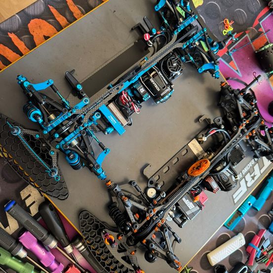 Ein zerlegter blauer und schwarzer RC-Wagen liegt auf einer grauen Unterlage. Verschiedene Werkzeuge und Teile sind darum verstreut, wobei einige Teile mit 'Team Associated' gekennzeichnet sind.