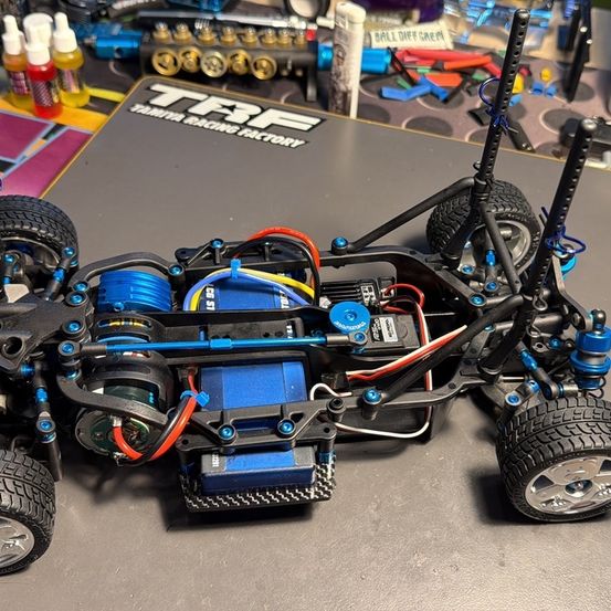 Ein schwarz-blaues Tamiya-Rennauto steht auf einer grauen Oberfläche. Das Auto hat sichtbare elektronische Komponenten, einschließlich einer Batterie und Drähte. Verschiedene Werkzeuge und Flaschen sind dahinter verstreut.