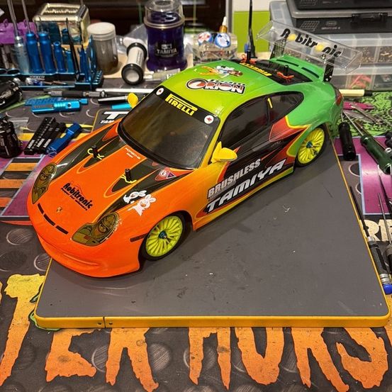 Ein leuchtend orange-grüner Tamiya GT3 RS Sportwagen-Modell wird auf einem grauen Untergrund ausgestellt, umgeben von verschiedenen Werkzeugen und Behältern auf einem Tisch.