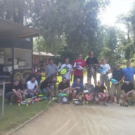 Eine Gruppe von Menschen posiert für ein Foto im Freien mit ihren ferngesteuerten Autos auf einer Schotterpiste, einige sitzen, andere stehen, unter einem Vordach und in der Nähe von Bäumen.