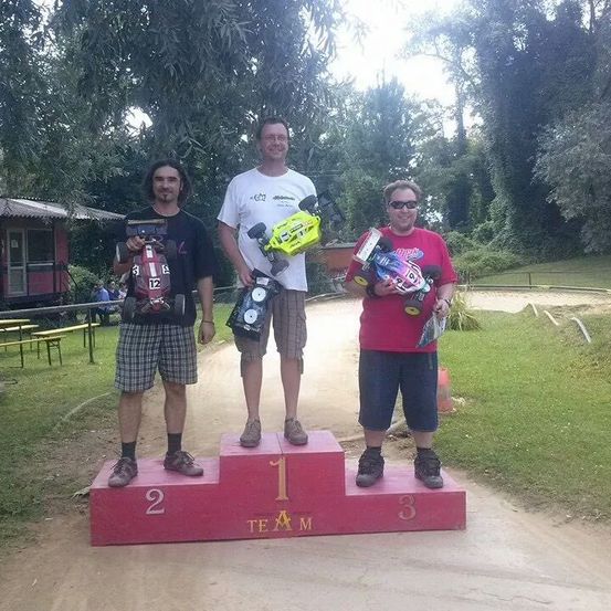 Drei Männer stehen auf einem roten Podest mit den Nummern 1, 2 und 3. Sie halten ferngesteuerte Autos. Dahinter steht ein kleines Gebäude, Bänke und Grünfläche.