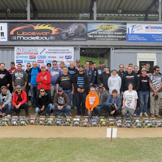 Eine Gruppe von Menschen posiert für ein Foto vor einem Gebäude mit einem Banner. Sie tragen Jacken, Mützen und Sneakers. Mehrere ferngesteuerte Autos stehen auf dem Boden.