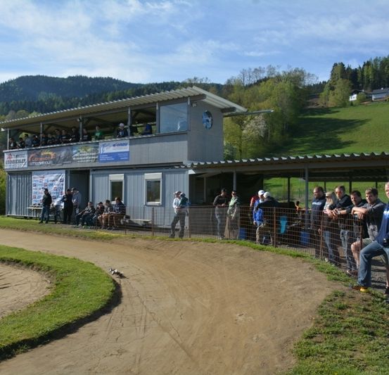 Eine Gruppe von Menschen beobachtet ein Dirt-Track-Event. Zuschauer stehen an einem Zaun, während andere auf einer Bank sitzen. Im Hintergrund befindet sich ein Gebäude mit Bannern und einem Aussichtsbereich.