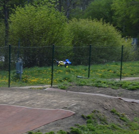 Eine Erdbahn mit Zaun und Büschen, eine Person in oranger Weste beobachtet ein Spielzeugauto in der Luft.