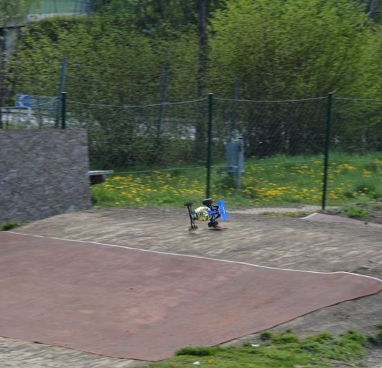 Eine Dirt-Rampe mit einem Spielzeugauto darauf, umgeben von Gras und Büschen, eingezäunt von einem grünen Zaun.