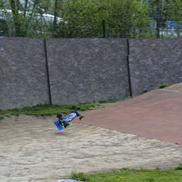 Ein Skateboard ist in der Luft über einer Dirt-Rampe mit einer Steinmauer dahinter, umgeben von Grün und einem Zaun.
