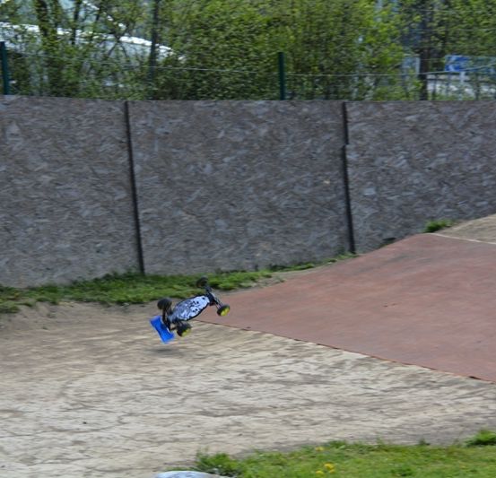 Ein Skateboard ist in der Luft über einer Dirt-Rampe mit einer Steinmauer dahinter, umgeben von Grün und einem Zaun.