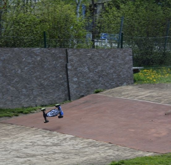 Ein blaues Skateboard mit einem schwarzen Rad ist in der Luft in einem Außenbereich, in der Nähe einer Betonmauer und Grünflächen.