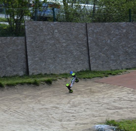 Ein ferngesteuertes Skateboard führt eine Stunt auf einer Erdbahn aus, mit einer Steinmauer und Grünpflanzen im Hintergrund.
