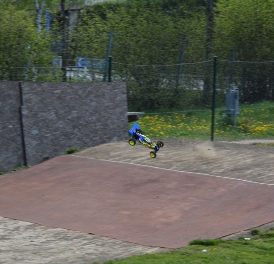 Ein blaues Spielzeugauto schwebt über einer Erdbahn, umgeben von einer Steinmauer und einem Metallzaun mit Bäumen und Gras im Hintergrund.