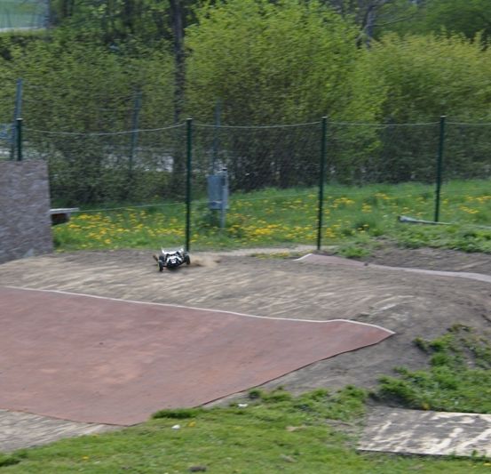Ein ferngesteuertes Auto befindet sich auf einer Erdrennstrecke mit Zaun, Wand und Bäumen im Hintergrund.