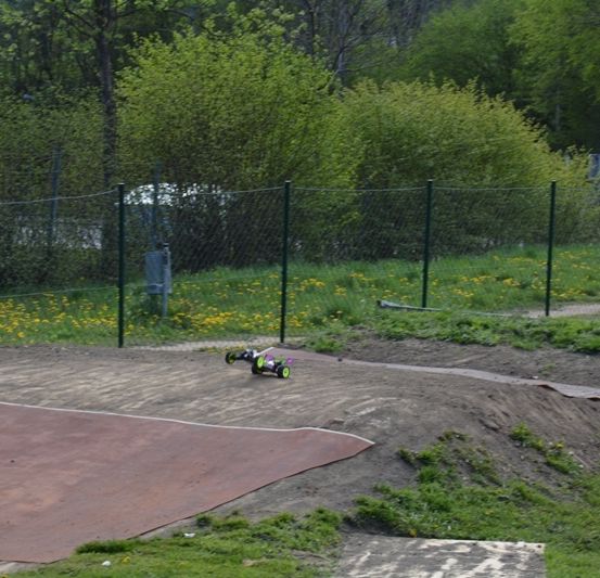 Ein ferngesteuertes Auto ist auf einer Erdbahn mit einer Rampe und einem Zaun, in der Nähe eines Grasbereichs mit Büschen und einem Schild.