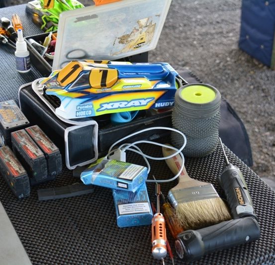 Eine Sammlung von Werkzeugen und Materialien liegt auf einem Tisch, darunter ein Werkzeugkasten, ein Pinsel, Schraubendreher und verschiedene elektronische Komponenten. Die Umgebung scheint im Freien zu sein.