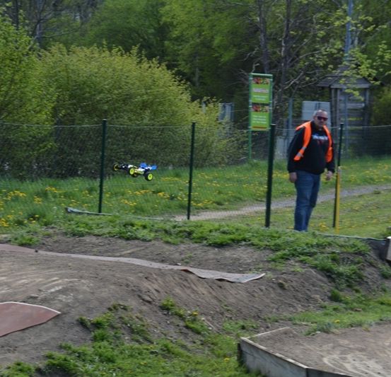 Ein Mann in einer orangefarbenen Weste steht in der Nähe einer Erdstrecke mit einem ferngesteuerten Auto darauf. Die Strecke ist eingezäunt, und es gibt Büsche und Bäume im Hintergrund.